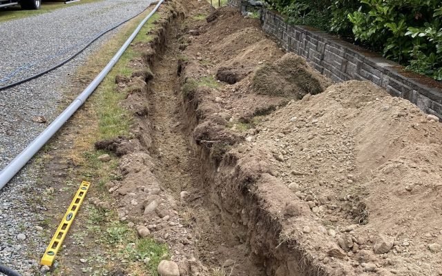 Open utility trench running alongside a gravel driveway, with grey conduit laid ready for installation and a stone block retaining wall to the right.