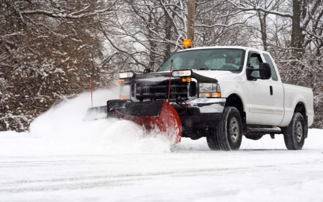 Pickup truck with a front plow blade clearing snow from a road, throwing a spray of snow